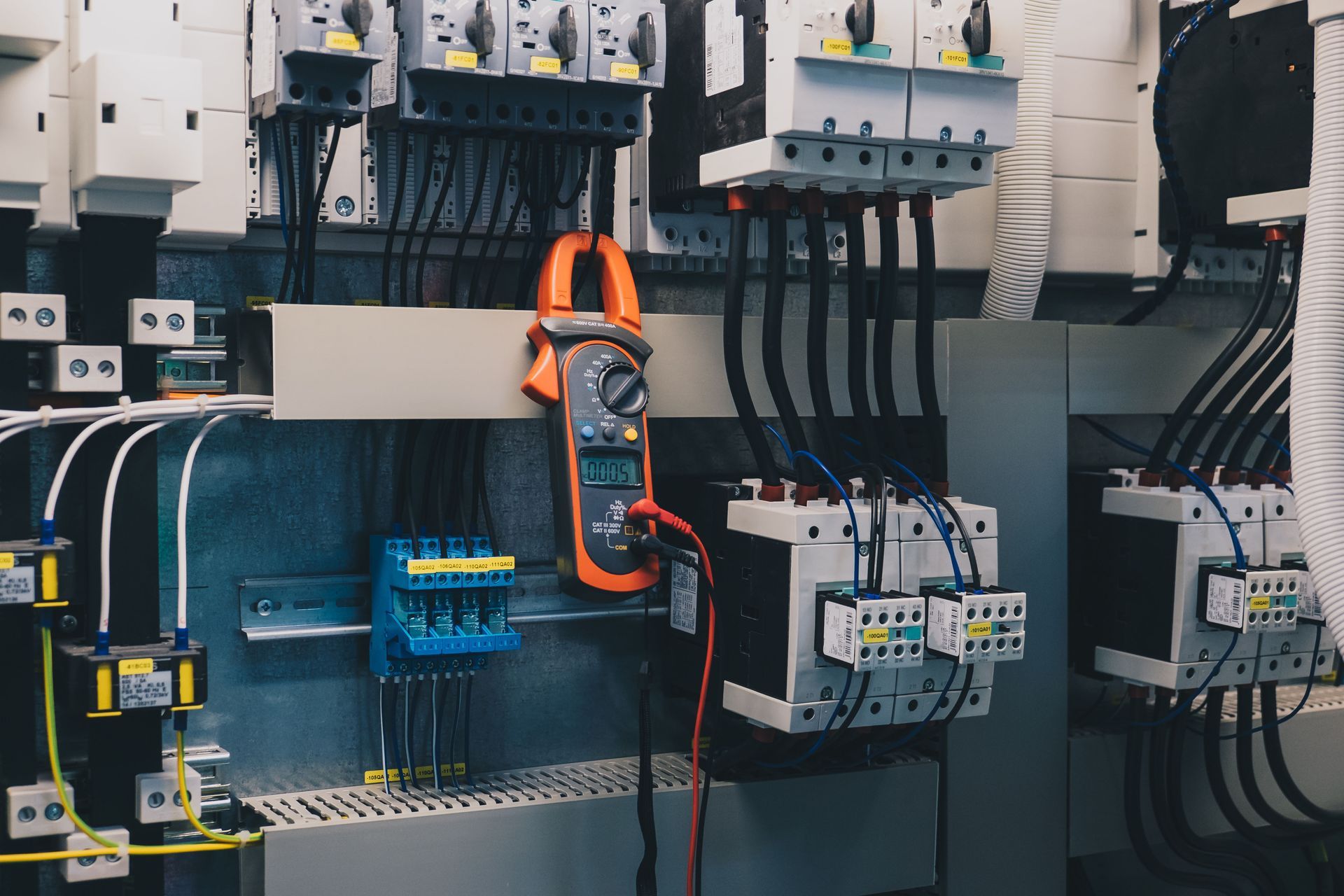 Electrical control panel with wiring and circuit breakers, including an orange multimeter hanging in the center.