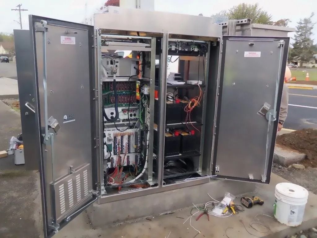 Open electrical control cabinet with exposed wiring and components outdoors beside a roadway.