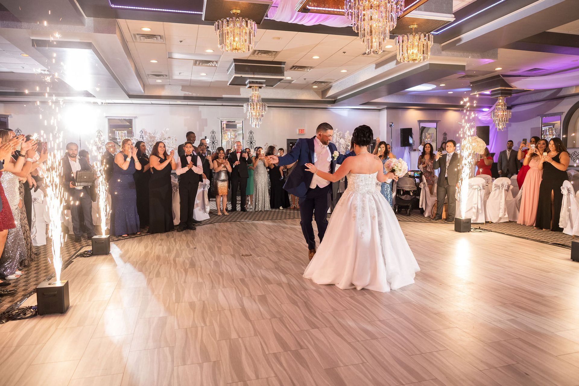 Newlyweds dance under sparklers at their wedding reception in a decorated ballroom. Guests applaud from the perimeter.