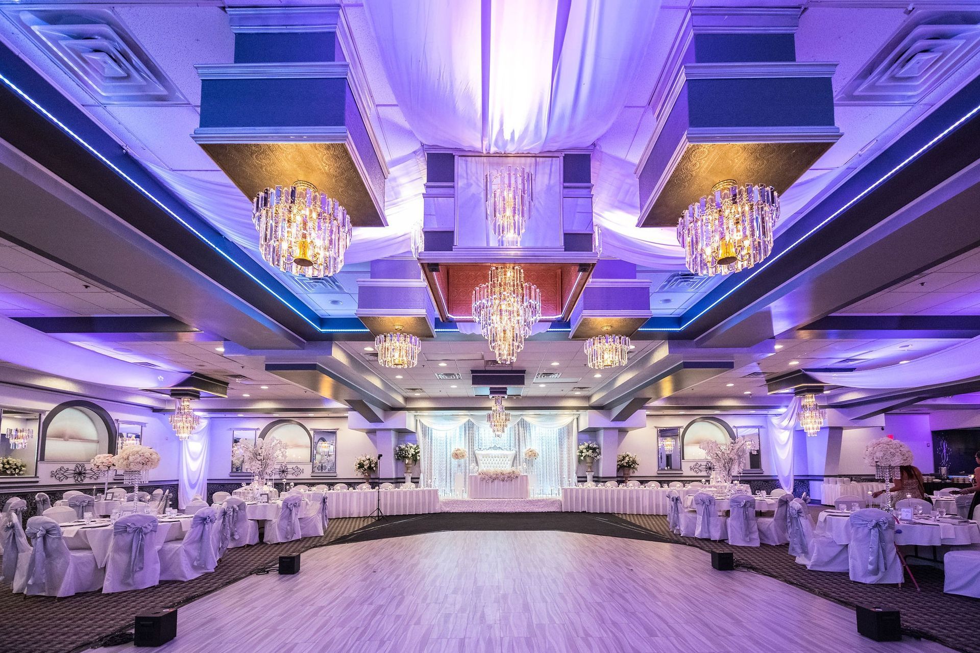 Elegant ballroom set for an event, with round tables, white linens, and ornate chandeliers. Purple and blue ambient lighting.