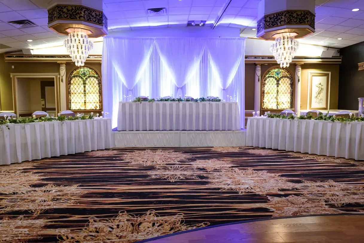 A ballroom with a draped stage, long tables, ornate lighting, and a patterned carpet.