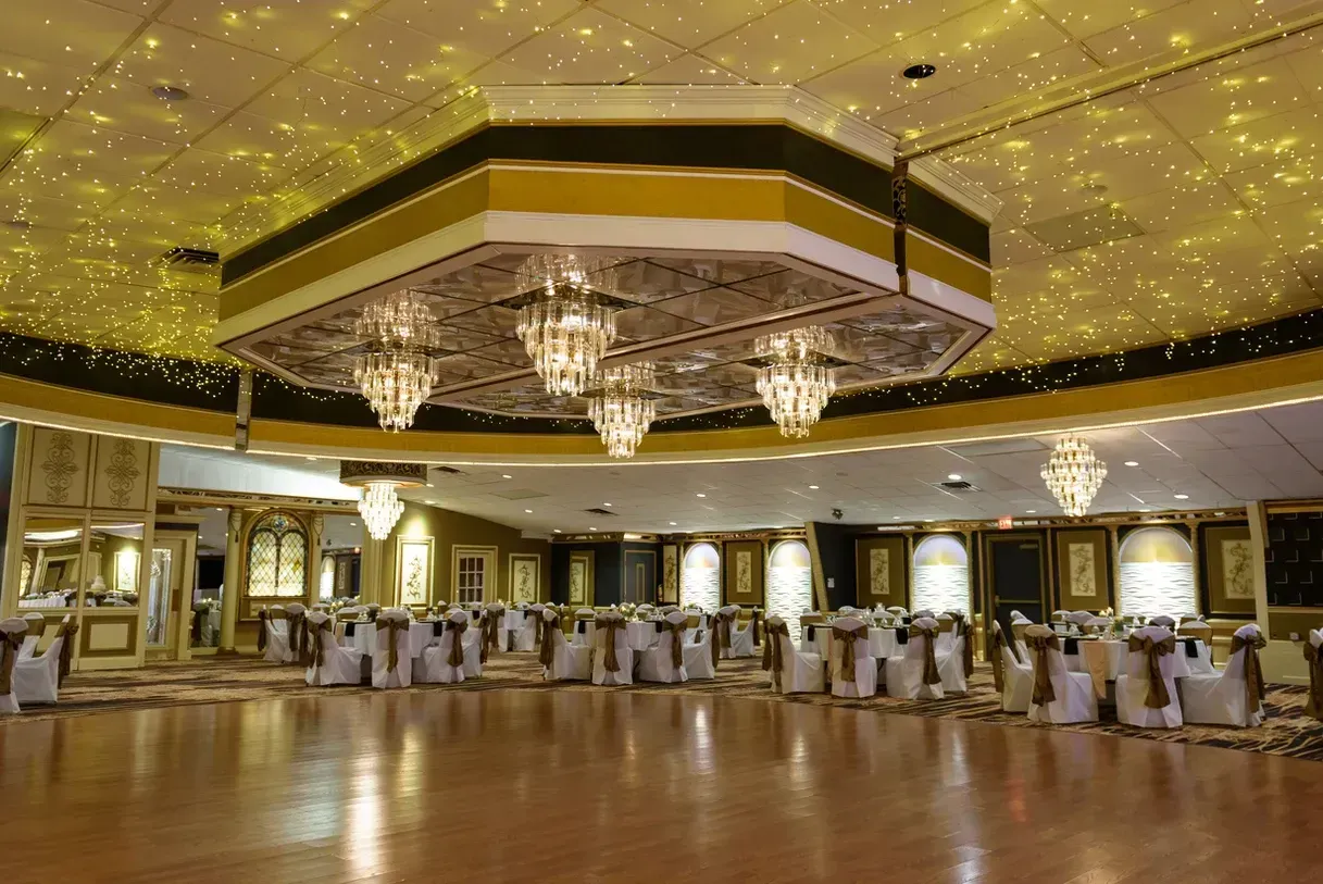 A large ballroom with round tables set for an event. Gold and white decor, ornate chandeliers, and a starry ceiling.