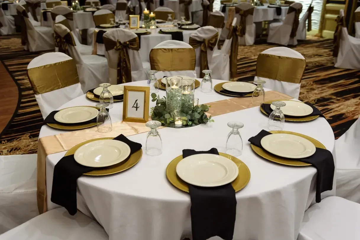 Round table set for a formal event, with gold chargers, black napkins, and gold-accented white chairs in a ballroom setting.