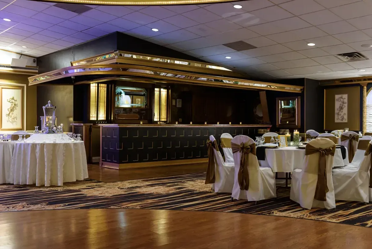 Elegant ballroom with round tables, chairs adorned with brown sashes, and a large, ornate bar in the background.