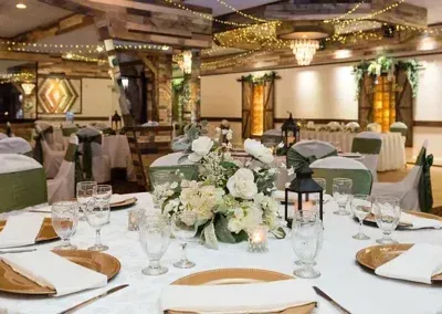 A wedding reception setup with round tables, white tablecloths, gold chargers, and floral centerpieces. The room is decorated in neutral tones with string lights.