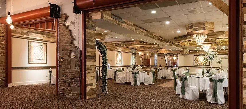 Interior of a decorated event hall with tables set for a gathering. Tables have white tablecloths with green sashes on chairs. Stone and wood decor.