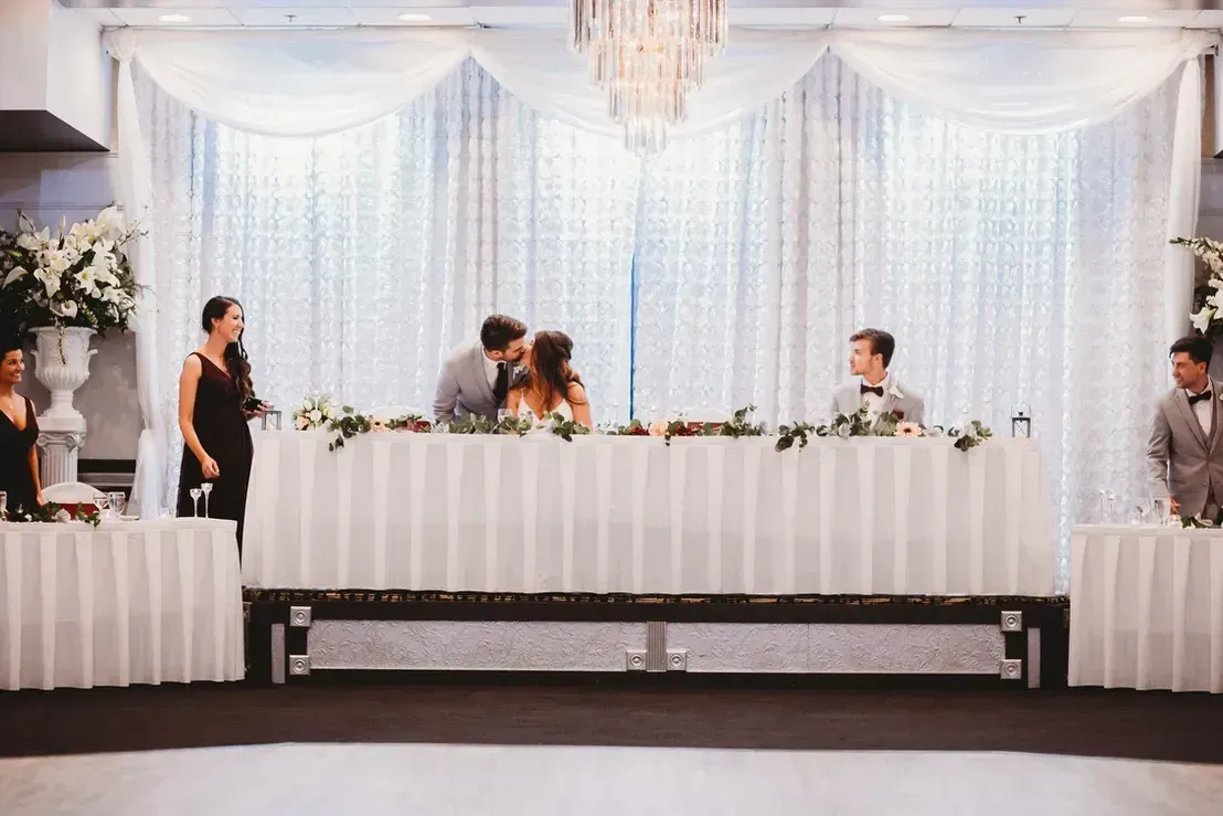 Bride and groom kiss at a reception table.  A floral garland adorns the table, and two attendants flank the couple.