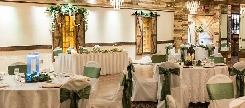 A wedding reception hall with round tables set for guests. White tablecloths, green chair sashes, and floral centerpieces decorate the room.