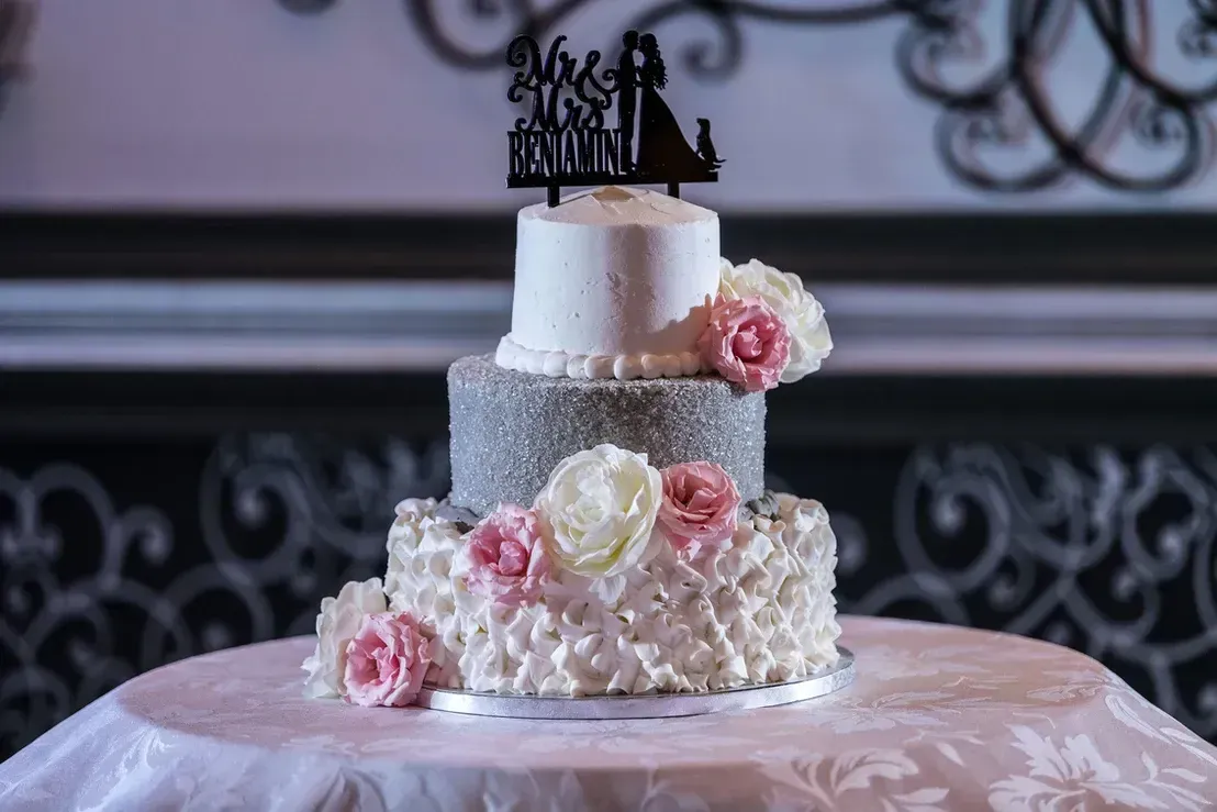 Three-tiered wedding cake with white, silver, and textured frosting, decorated with pink and white flowers, and a bride and groom cake topper.