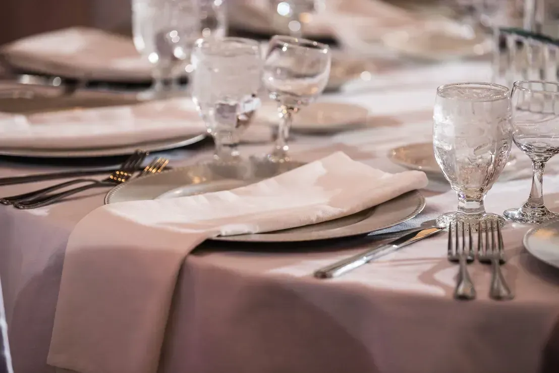 A table set for a formal event, with plates, silverware, water glasses, and a folded white napkin on a pink tablecloth.