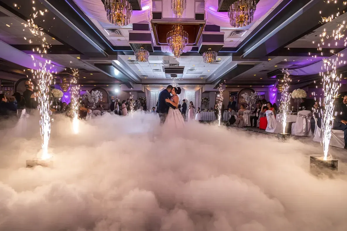 Couple dancing in a ballroom surrounded by fog and sparklers during a wedding celebration. Elegant setting with guests seated at tables.
