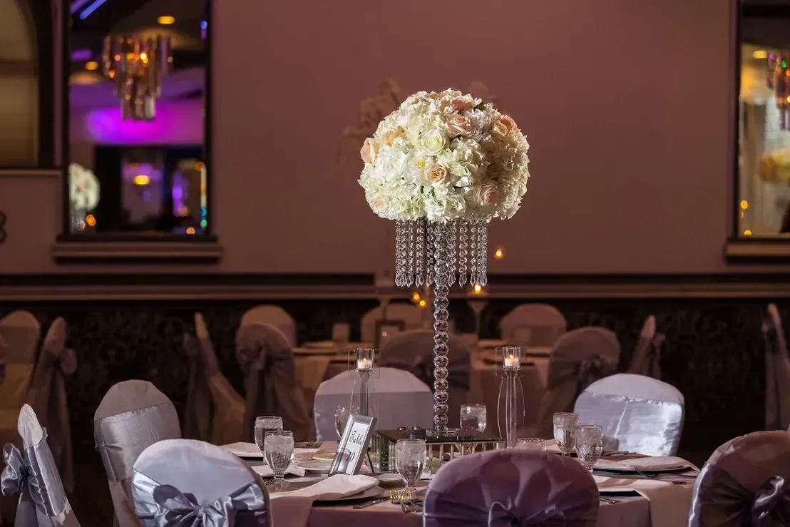 A decorated table with a tall floral centerpiece and covered chairs, set in a formal event space.