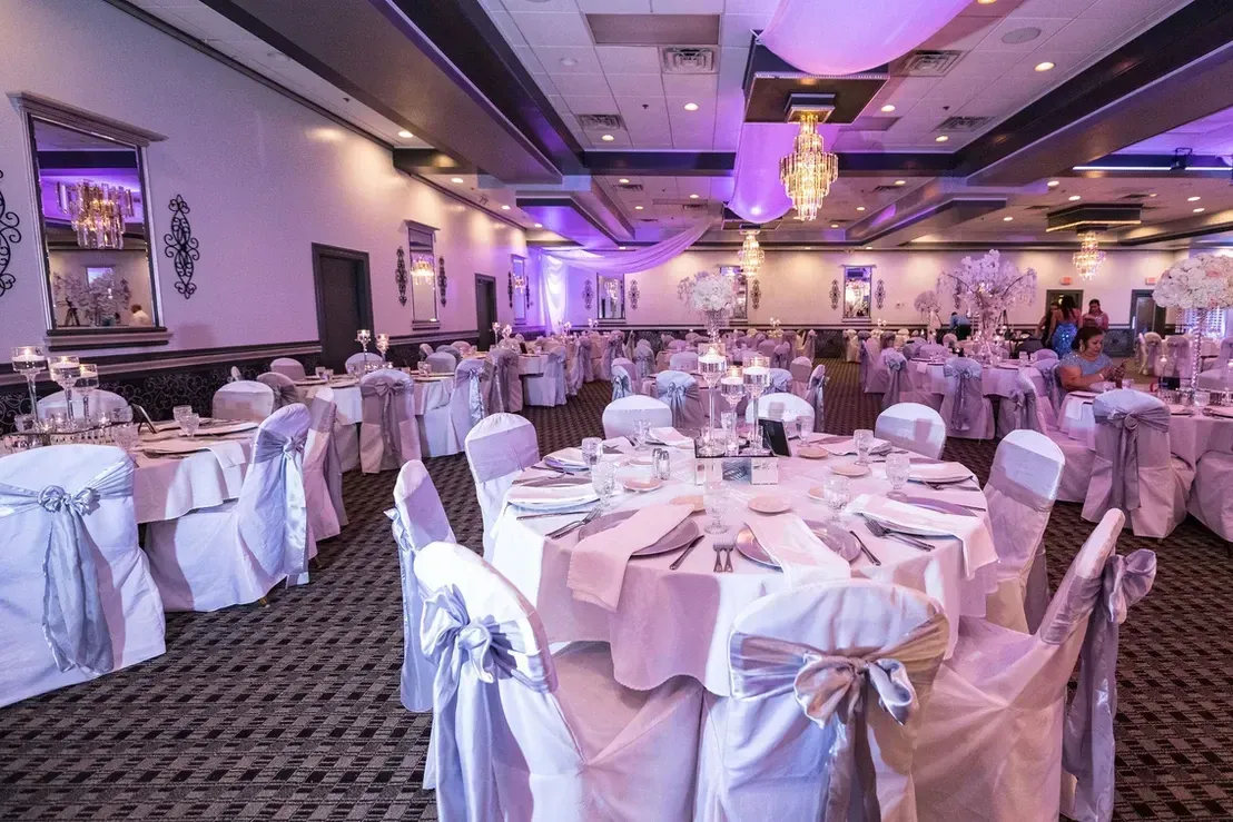 A banquet hall with round tables set for a formal event; tables have white linens, chair covers, and centerpieces, with purple lighting.