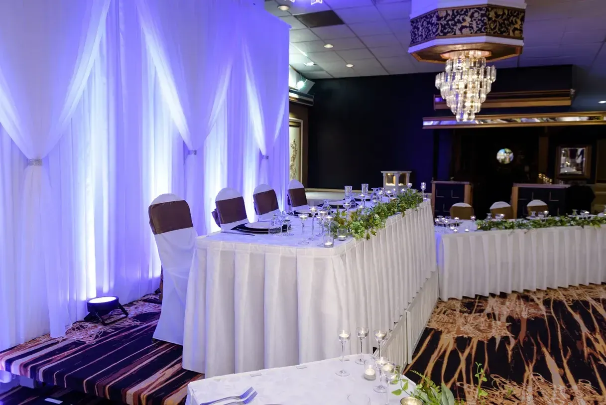 Wedding reception with a long table set for the wedding party; white table linens and floral arrangements; blue uplighting, ornate chandelier.