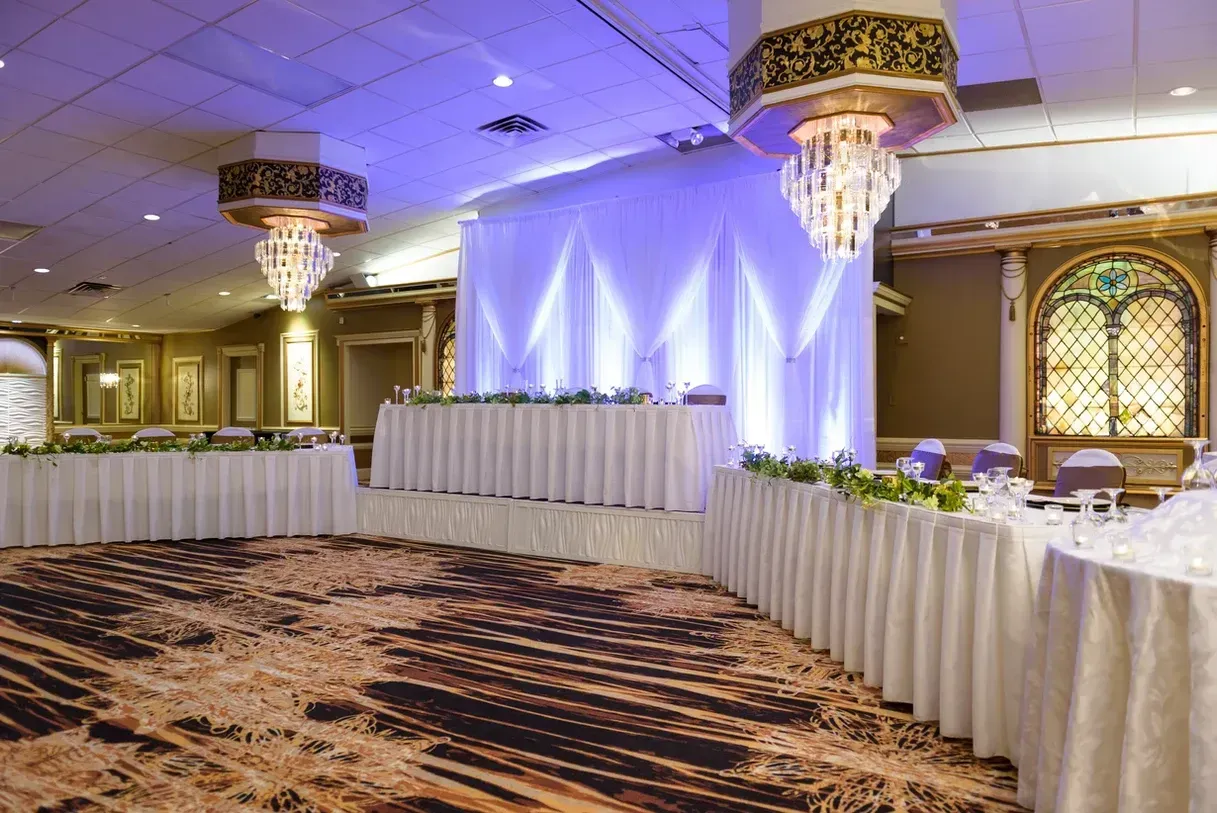 A wedding reception hall with white draped backdrop, long tables, floral centerpieces, and a patterned dance floor. Two ornate chandeliers hang from the ceiling.