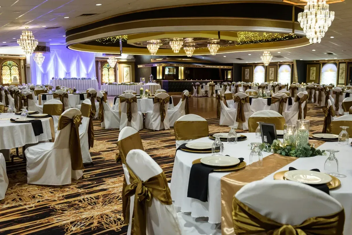 Elegant ballroom set for an event with round tables covered in white cloths, gold accents, and ornate chandeliers.