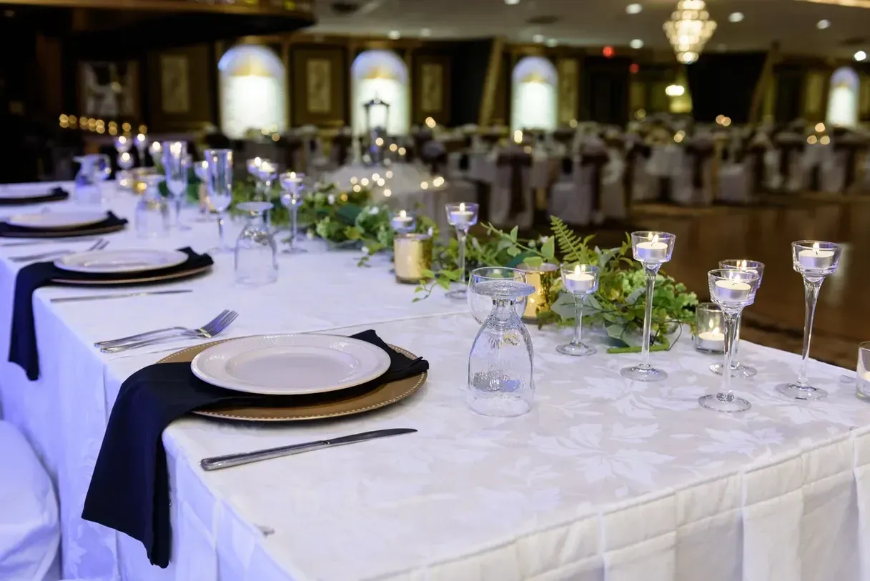 Long table set for a formal event, with plates, silverware, candles, and greenery.  Room has dark chairs and decorative arches.