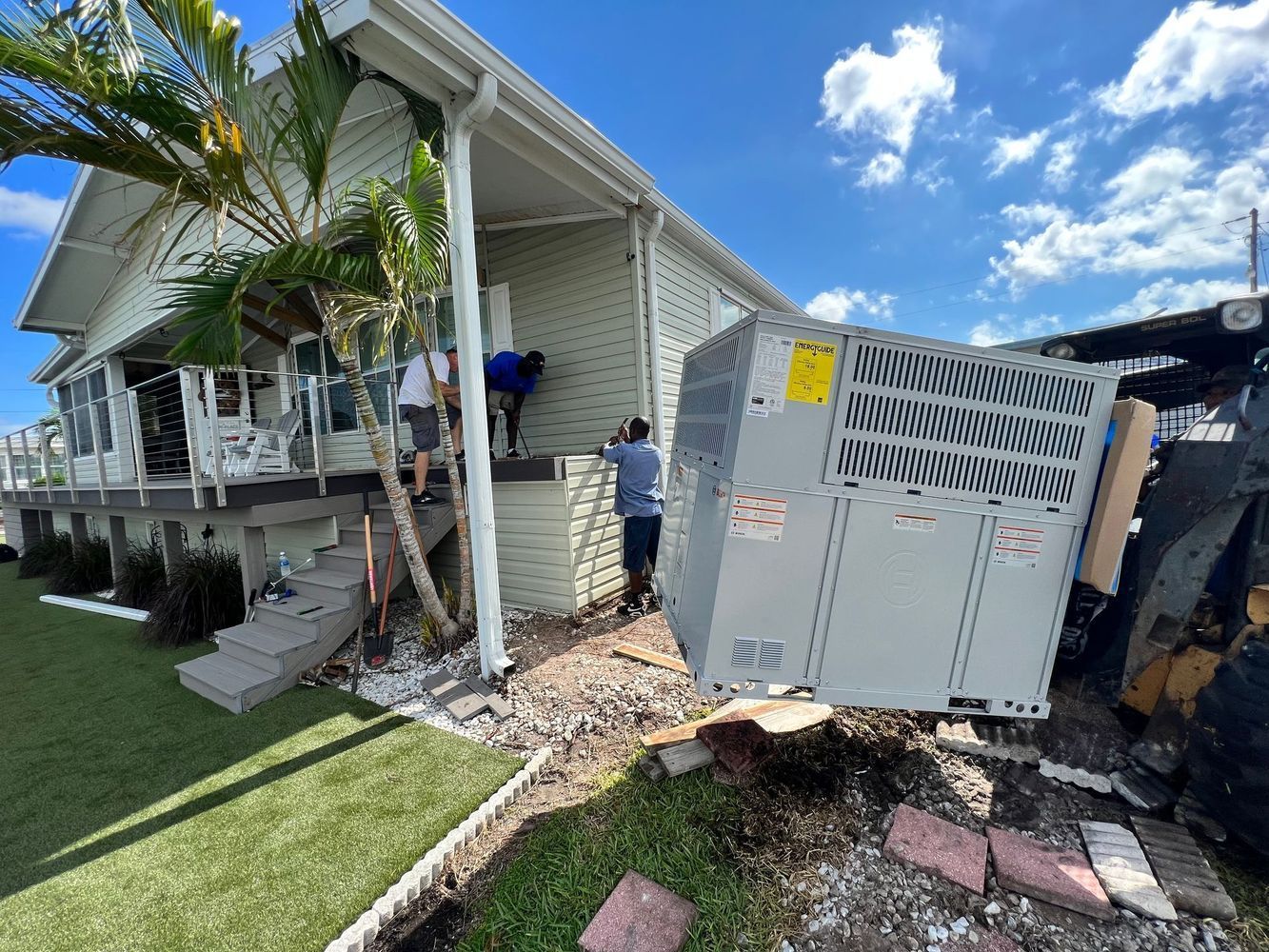 Workers installing a large air conditioning unit at a light green house under a blue sky.