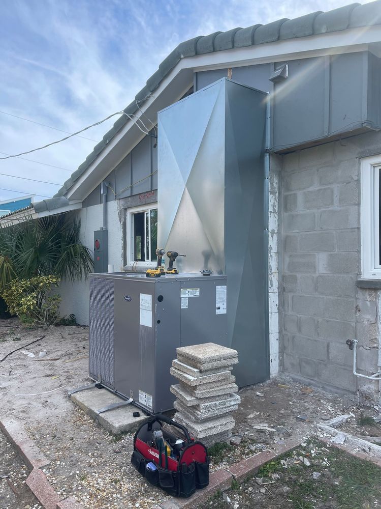 HVAC unit installed outside a house with a metal duct, tools and concrete blocks visible.
