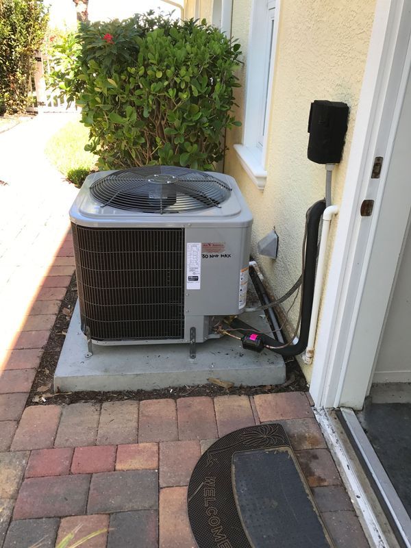 An air conditioning unit next to a building, on a concrete base, with brick pathway.