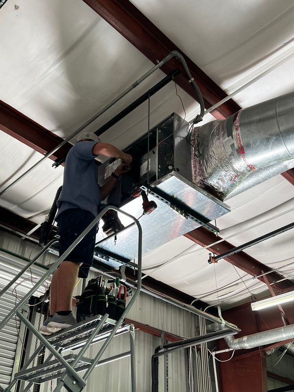 HVAC technician on a metal staircase installs ductwork in a building. He is using a power tool and wearing shorts.