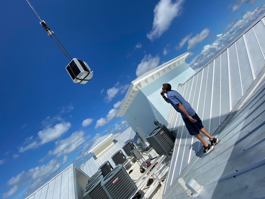 Man on rooftop observing HVAC unit being lifted by crane against a blue sky.