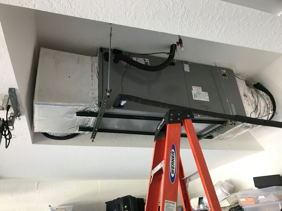 An air handler unit installed in a garage ceiling; a stepladder is set up below.
