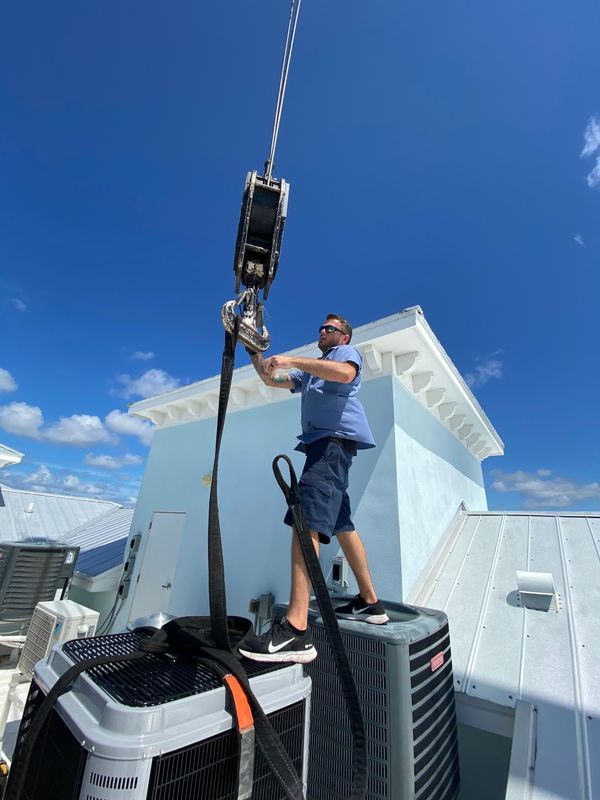 Man on rooftop secures HVAC unit to crane hook under blue sky.