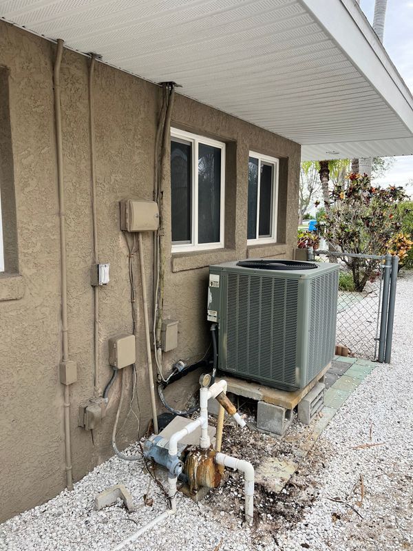 Air conditioning unit outside a stucco building with electrical conduits and drainage pipes.