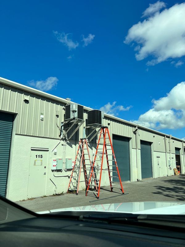 Exterior view of a building with roll-up doors, air conditioning units, and three orange ladders set up below.