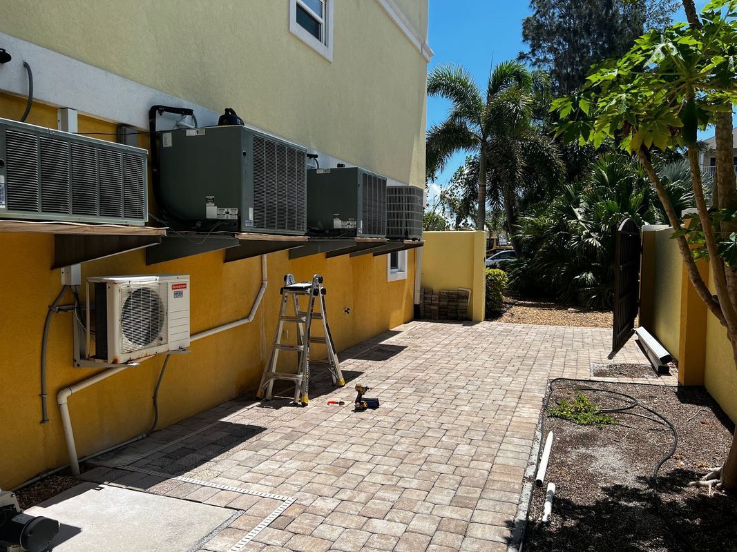 Air conditioning units mounted on a yellow building exterior, next to a paved patio.