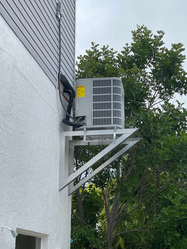AC unit mounted on a bracket attached to a gray and white building wall, near trees.