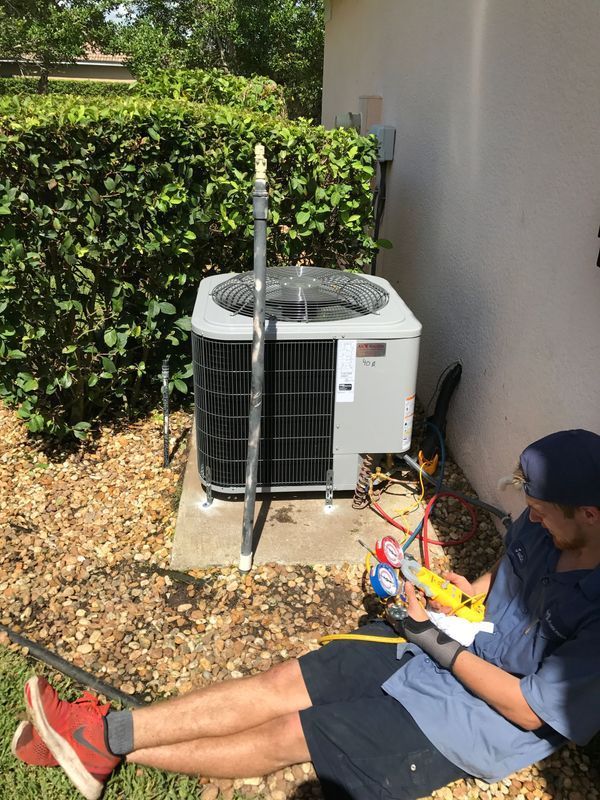 AC technician seated by an outdoor unit, checking gauges, and working on an HVAC system in a residential setting.