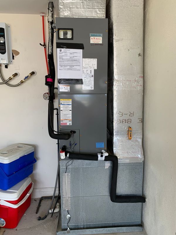 HVAC unit in a garage. Gray and silver components, with black insulated pipes and white walls.