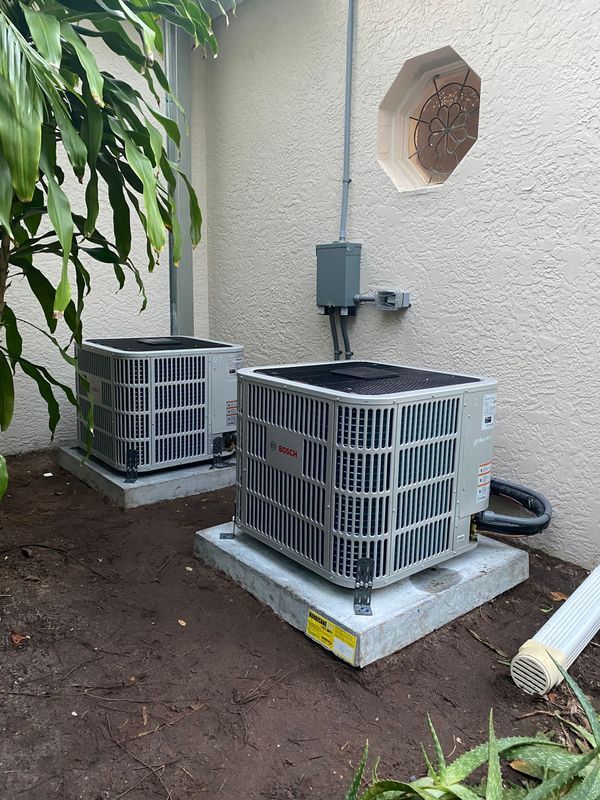 Two air conditioning units sit on concrete pads outside a beige building, with a wasp nest nearby.