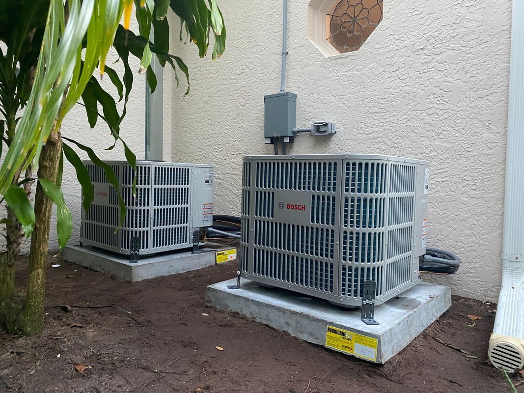 Two air conditioning units on concrete pads next to a building and greenery.