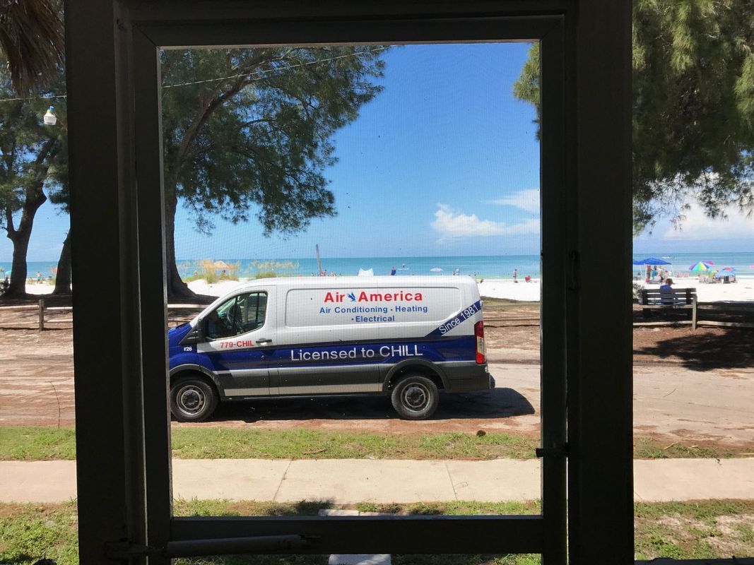 View through a window, a white van with blue lettering parked near a beach with blue sky and ocean.