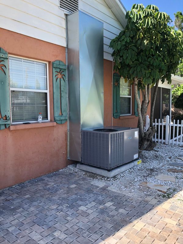 Air conditioning unit outside a peach-colored house with green shutters; metal ductwork is attached to the house.
