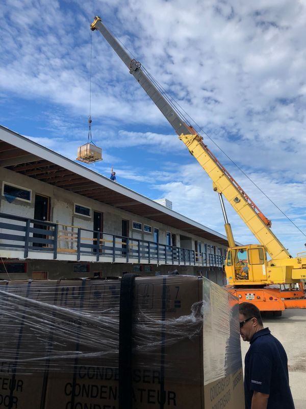 A crane lifting an object onto a building roof; boxes in the foreground. Man watches. Blue sky.