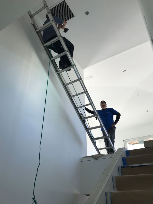 Two people on a ladder, one in an attic access. Another person stands on the stairs. Interior shot, white walls.