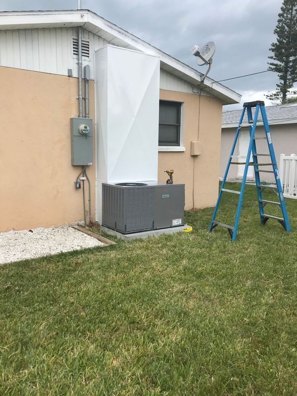 Air conditioner unit and white cover installed on a beige house with a ladder and green grass in a yard.