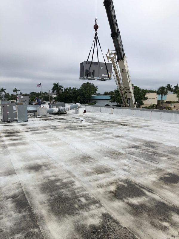 A crane lifting an air conditioning unit onto a flat commercial roof. Overcast day.