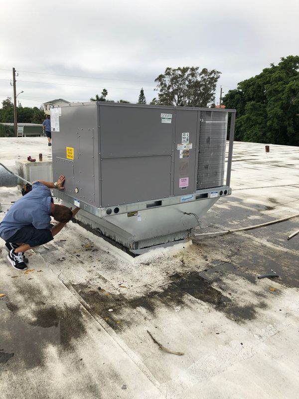 Two people working on a rooftop HVAC unit; gray unit on a white roof, overcast sky.