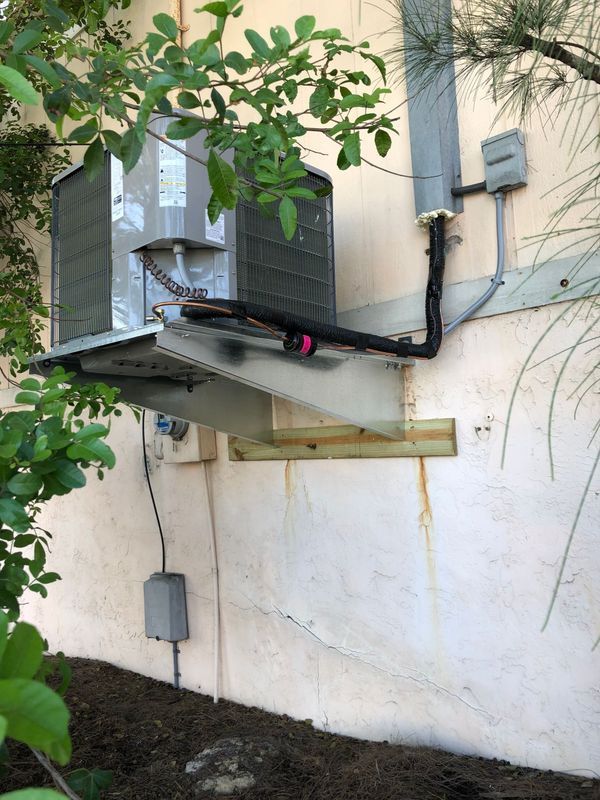 AC unit mounted on a building exterior with associated pipes, wiring, and supports. Beige wall, green foliage, and brown mulch.
