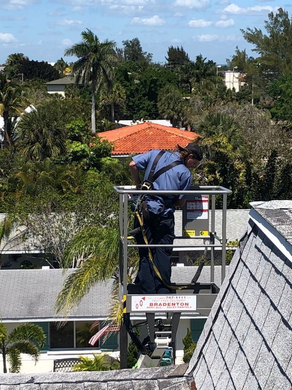 Person on lift inspecting a roof. Blue shirt, safety harness. Sunny day, residential setting.