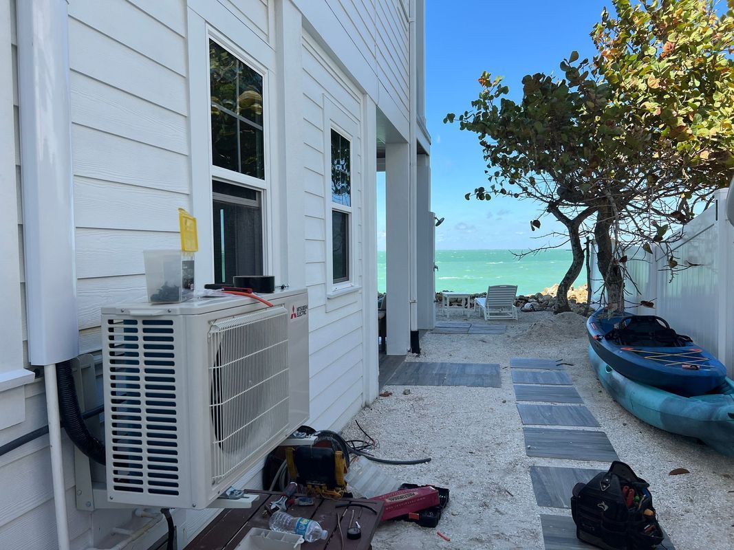 White building with an AC unit and tools, a path to the ocean with a kayak and tree, under a blue sky.