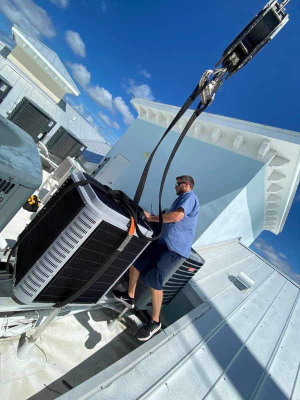 Man on rooftop securing an air conditioning unit being lifted by a crane against a blue sky.