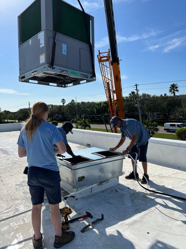 HVAC unit lifted by crane, being installed on a roof by three workers on a sunny day.