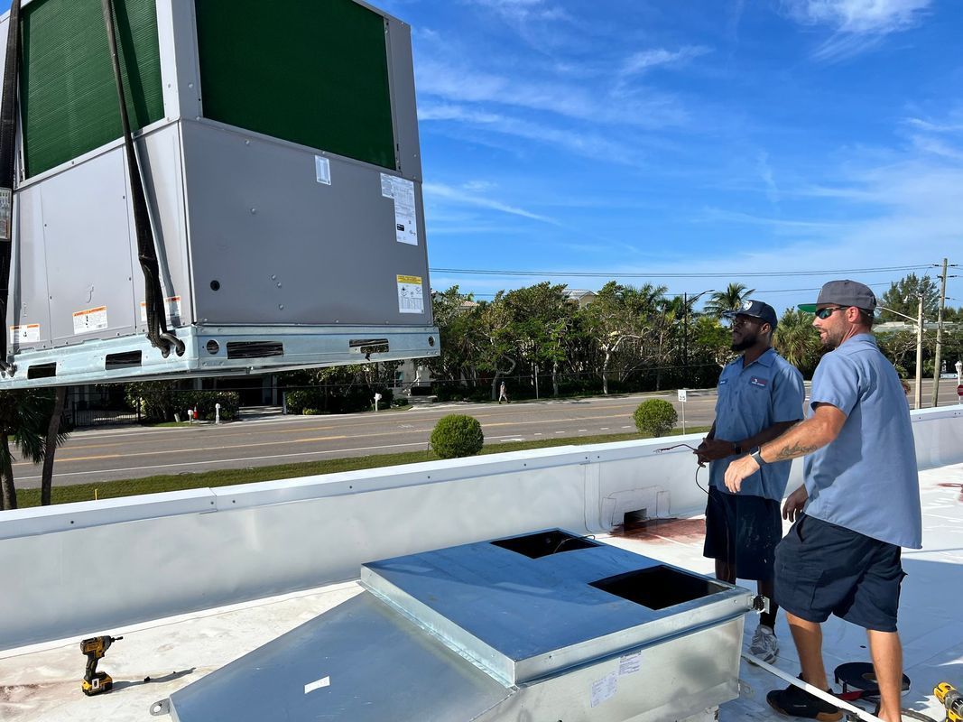 Two workers on a rooftop oversee the installation of an HVAC unit, under a blue sky.