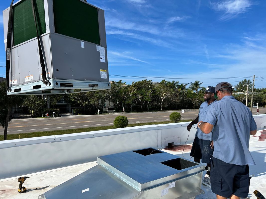 HVAC unit being lifted onto a roof by a crane; two workers watch, sunny day.